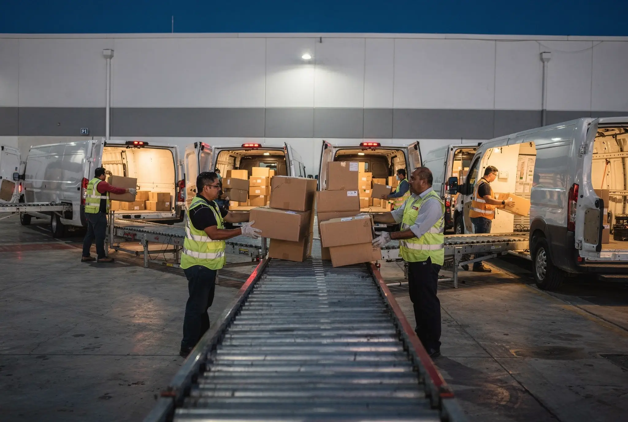 Trabajadores cargan paquetes de cartón desde una banda de rodillos a camionetas blancas de reparto en el andén de un centro de fulfillment al anochecer.