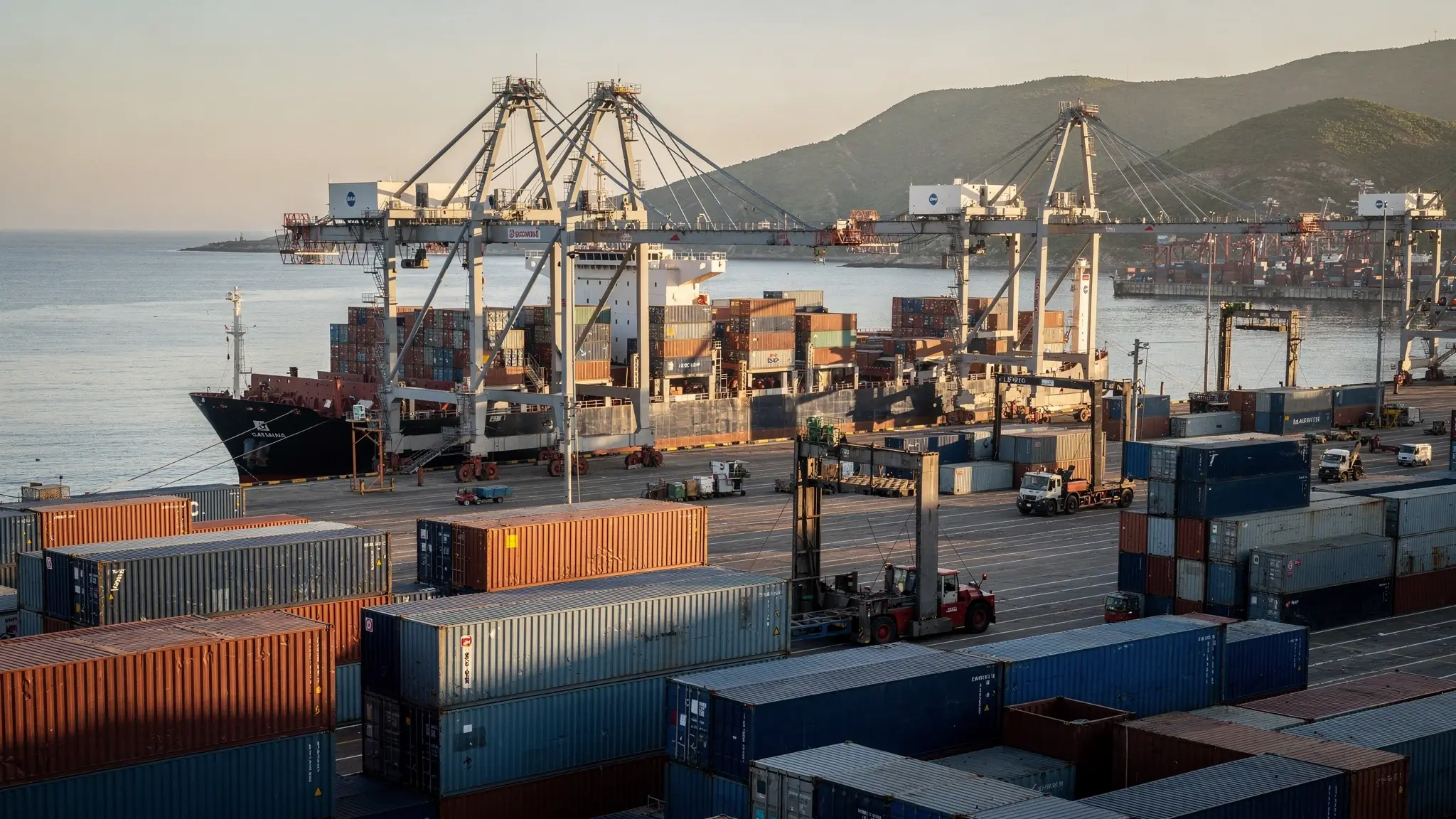 Pilas de contenedores y grúas de pórtico en un puerto del Pacífico durante la hora dorada, con cerros verdes alrededor de la bahía.